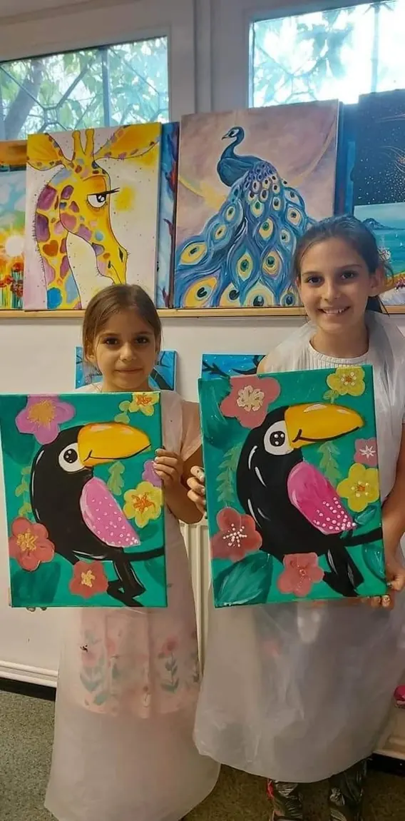 Dos niñas sostienen los tucanes que acaban de pintar, con más obras del taller al fondo.